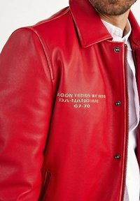 Veste en cuir rouge au toucher lisse, col pressionné et fermeture éclair argentée. Elle présente un texte brodé sur la partie gauche de la poitrine.