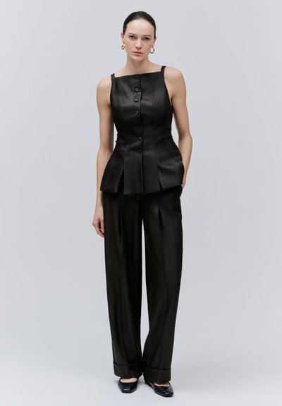 Vrouw in een mouwloze zwarte top met knopen en plooien, zwarte broek met wijde pijpen, zwarte puntige schoenen, staand tegen een effen grijze achtergrond.