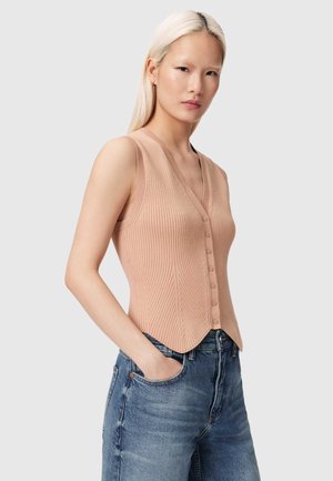 Jeune femme aux cheveux blonds raides portant un gilet beige côtelé sans manches à boutons et un jean bleu, debout les mains dans les poches.