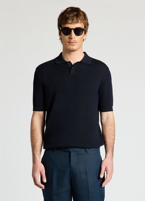 Homme portant un polo noir texturé à manches courtes, un pantalon foncé et des lunettes de soleil rondes noires, debout devant un fond clair et uni.