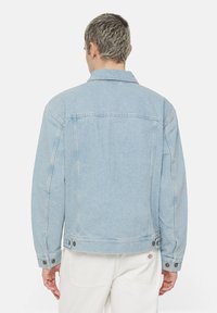 Ljusblå denimjacka med klassisk krage, knäppta ärmslut och sidofickor. Bakvy visar sömnadsdetaljer och slät textur.