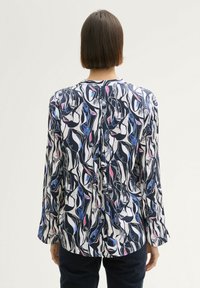 Blouse à manche longue en tissu fluide avec un motif abstrait aux teintes noires, bleues et roses. La vue de dos révèle un détail fendu subtil.