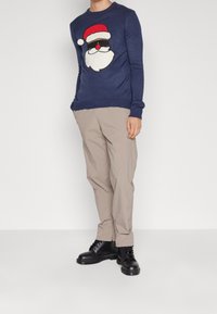 Navyblauer Pullover mit Rundhalsausschnitt, verziert mit einem großen Weihnachtsmann-Gesicht mit roter Mütze und Sonnenbrille. Beige Hose und schwarze Schuhe.