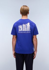 Blå bomulds T-shirt med en grafik af skiudstyr på ryggen, med mærkenavnet "Napapijri Geographic" i orange tekst.