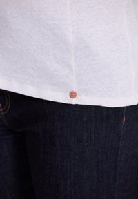 Witte katoenen t-shirt met een afgeronde zoom, voorzien van een klein koperen label nabij de zoom, gecombineerd met donkere denim jeans.