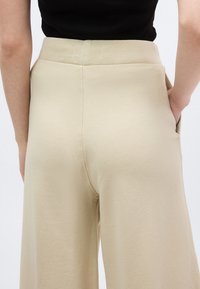 Άτομο που φοράει μπεζ παντελόνι με ψηλή μέση και φαρδιά πόδια, με τα χέρια στις πλαϊνές τσέπες, συνδυασμένο με μαύρο τοπ, εμφανιζόμενο από την πίσω πλευρά.