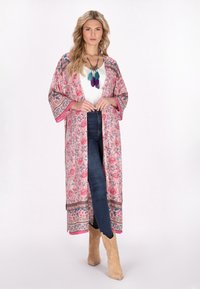 Kimono rosa con motivo floreale, maniche larghe e bordi a contrasto, abbinato a una maglietta bianca e jeans scuri, indossato con stivali marrone chiaro.