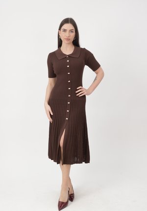 Vrouw staat met hand op heup, gekleed in een bruine geribbelde midi-jurk met knopen, korte mouwen en een kraag, gecombineerd met bordeauxrode hakken.