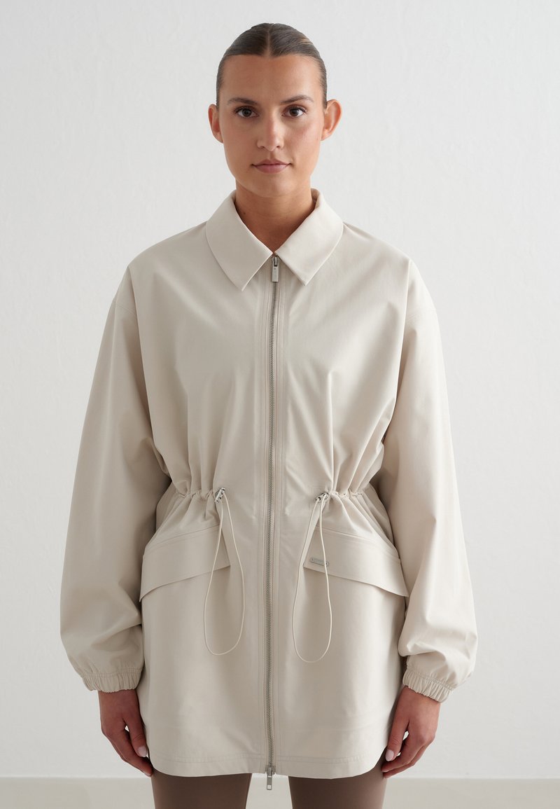 Femme portant une veste beige à fermeture éclair avec cordons à la taille et poignets élastiques, debout devant un fond blanc uni.