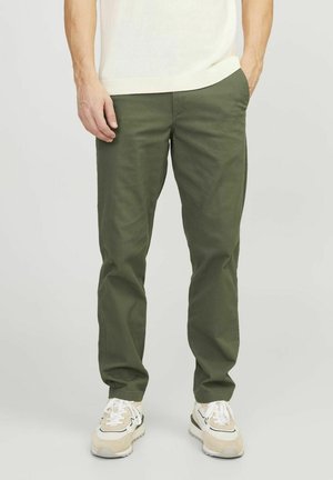 Man draagt olijfgroene casual broek, witte sneakers en een witte top met korte mouwen, staat met handen deels in de zakken.