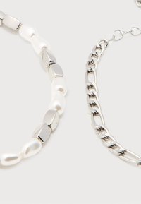 Δύο βραχιόλια δίπλα δίπλα: το ένα με λευκές πέρλες και ασημένιες γεωμετρικές χάντρες, το άλλο μια ασημένια αλυσίδα με ενωμένους κρίκους.