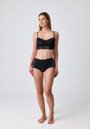 Vrouw die blootsvoets staat, gekleed in een zwarte kanten bralette en hoge tailleonderbroek met kanten panelen, licht naar rechts kijkend tegen een effen achtergrond.