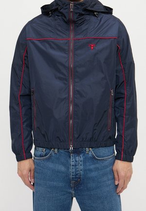 Veste zippée bleu marine avec passepoil rouge et petit logo rouge sur la poitrine, portée avec un jean bleu, mains détendues le long du corps.