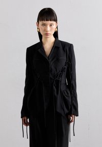 Schwarzer taillierter Blazer mit strukturierter Silhouette, der Schnürungen an den Seiten und vorne aufweist, hergestellt aus einem glatten Stoff mit matter Oberfläche.