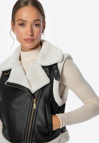Chaleco de imitación de cuero negro con cuello de borreguito blanco, cierre frontal con cremallera y bolsillos laterales. Presenta herrajes dorados y un diseño ajustado.