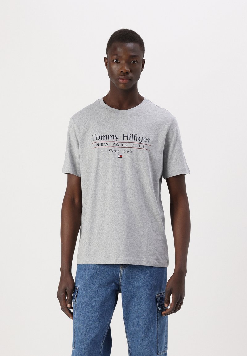 Grå t-shirt med korte ærmer og rund halsudskæring, med teksten "Tommy Hilfiger," "New York City," og "Since 1985" trykt i mørkeblå og rød.