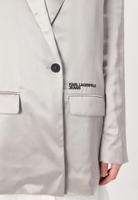 Lys grå blazer med en glatt tekstur, med en enkel knappåpning, to frontlommer og brodert "KARL LAGERFELD JEANS"-logo.