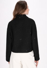 Sort tekstureret jakke med glitrende detaljer, der har krave, lange ærmer og et cropped design. Overfladen har et rutenetmønster.
