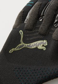 Gant de sport noir avec un tissu texturé, des accents de prise en caoutchouc et un logo de chat réfléchissant vert, avec des détails en rayures bleues au niveau du poignet.