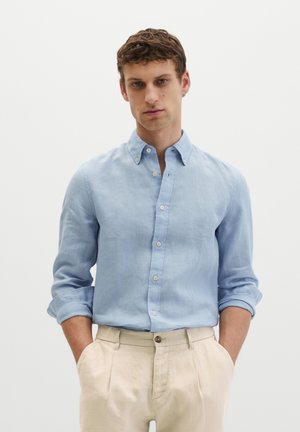 Junger Mann mit kurzen lockigen Haaren trägt ein hellblaues langärmeliges Hemd und beige Hose, Hände in den Taschen, neutraler Hintergrund.