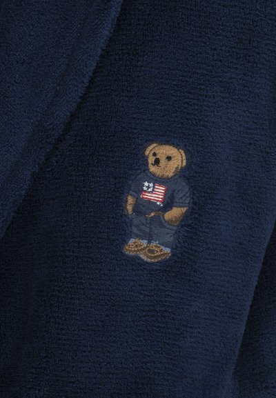 Námornícky modrá fleece látka s malým, vyšitým medveďom, ktorý má na sebe tričko s dizajnom americkej vlajky a džínsy.