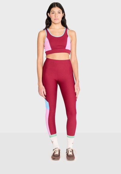 Femme debout, face à l'avant, portant des leggings bordeaux et un crop top assorti avec des blocs de couleur rose et bleu, ainsi que des baskets avec des chaussettes blanches.