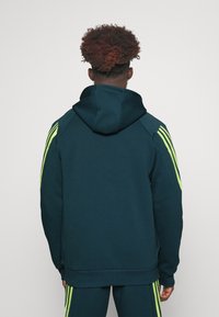 Kapućna majica tamno tirkizne boje s kontrastnim neonsko zelenim prugama duž ramena. Izrađena je od mekane tkanine, s opuštenim krojem.