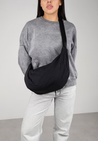 Bolso bandolera negro con una tela suave y texturizada, forma curva y una sola correa ajustable, llevado sobre un suéter gris y jeans claros.