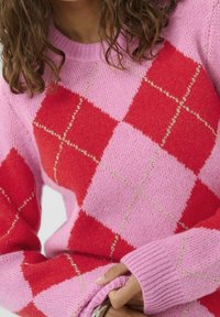 Personne portant un pull à motif losanges rose et rouge avec des accents de fil doré, tenant l'ourlet du pull d'une main.