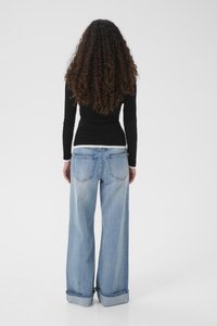 Donna con capelli lunghi e ricci vista di spalle, indossa una maglia nera a maniche lunghe e jeans larghi azzurri con orli risvoltati.
