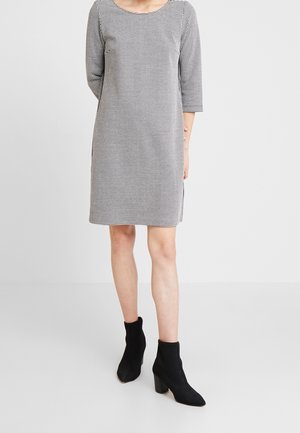 Femme portant une robe à carreaux noire et blanche longueur genou avec des manches trois-quarts et des bottines noires à talons, debout sur un fond uni.