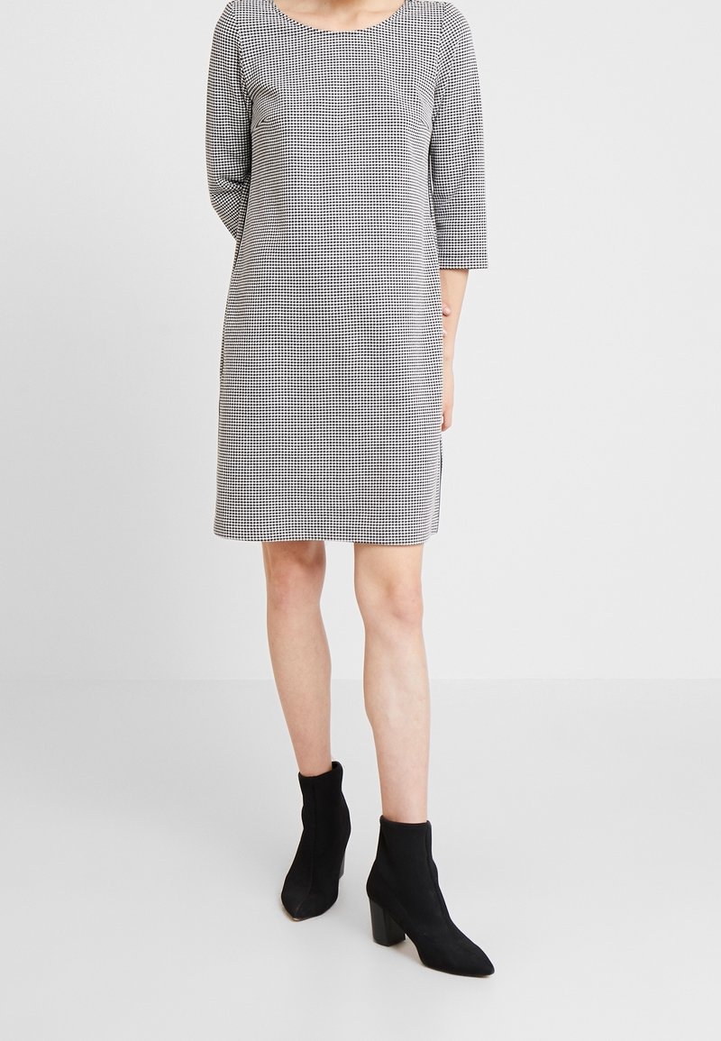 Femme portant une robe à carreaux noire et blanche longueur genou avec des manches trois-quarts et des bottines noires à talons, debout sur un fond uni.