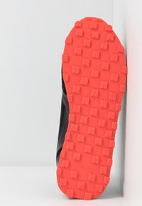 Chaussure noire avec une semelle orange vif présentant des crampons carrés en relief dans un motif répétitif, positionnée sur un fond blanc.