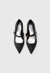 Zapatos en satén negro con punta afilada y un forro interior plateado, además de una correa estrecha. Presenta un detalle metálico decorativo cerca de la correa.