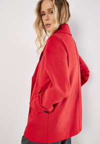 Punainen villasekoitteinen takki, jossa on rakenteellinen muotoilu, lovikaulus, kaksi sivutaskua ja nappikoristeita, esitelty sivukulmalta.