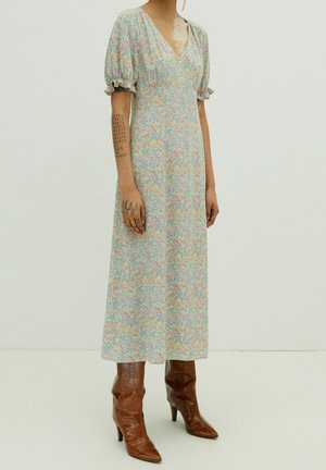 Personne portant une robe midi à fleurs pastel avec des manches ballon et des bottines marron à talons moyens pointues, se tenant contre un fond uni clair.