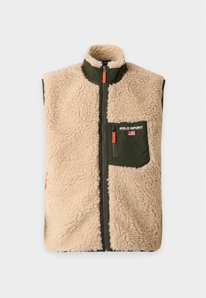 Homme portant un gilet en polaire beige avec une poche verte, une chemise manches longues bleu marine, un jean bleu clair et des baskets vertes avec des lacets rouges, debout avec la main dans la poche.