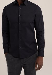 Muž v těsném černém košili s dlouhými rukávy a tmavě šedých kalhotách, s rukama částečně v kapsách, na béžovém pozadí.