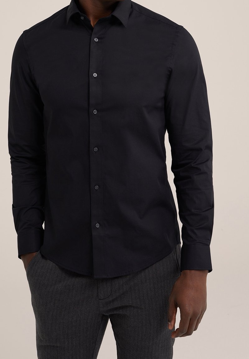 Muž v těsném černém košili s dlouhými rukávy a tmavě šedých kalhotách, s rukama částečně v kapsách, na béžovém pozadí.