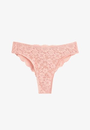 Kvinde med langt krøllet hår iført en hvid ribbet tanktop og lyserødt blonder højbenet undertøj, stående barfodet mod en ensfarvet baggrund.