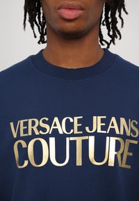 Sweatshirt bleu marine en coton avec un col côtelé, comportant un texte doré métallisé "VERSACE JEANS COUTURE" sur la poitrine.