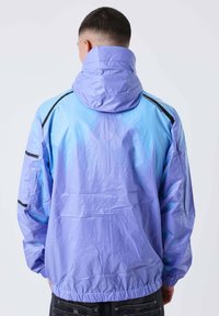 Uomo di spalle che indossa un windbreaker con cappuccio di colore azzurro chiaro e viola, con dettagli neri e polsini elasticizzati.