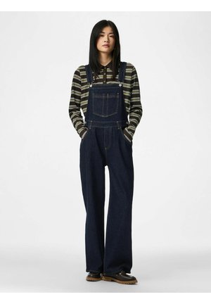 Femme debout portant une salopette en denim bleu foncé, un polo rayé à manches longues et des chaussures noires avec des semelles marron sur un fond blanc.