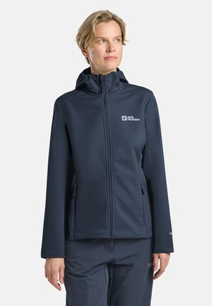 Vrouw met kort blond haar, gekleed in een donkerblauwe Jack Wolfskin jas en broek, kijkt iets naar links tegen een effen achtergrond.
