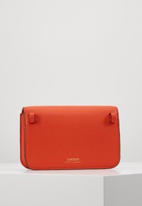 Pomarańczowa skórzana torebka o fakturowanej powierzchni, wyposażona w magnetyczne zapięcie i tłoczone złote logo. Kompaktowy, prostokątny kształt.