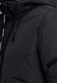 Svart vadderad jacka med hög krage, justerbar dragsko-huv hood och sidoficka med dragkedja. Har kontrasterande sömmar och logodetalj.