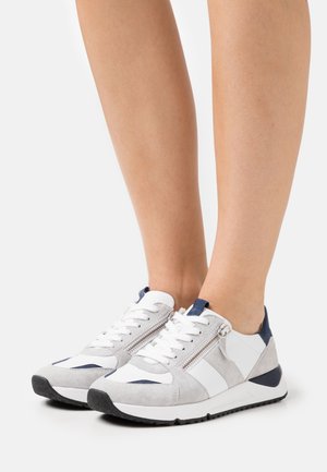 Sportovní tenisky v bílé barvě, šedé semišové a námořnické akcenty; mají šněrování v přední části a boční zip. Silná černá gumová podešev.