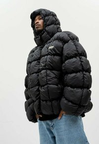 Doudoune noire avec capuche, présentant un design matelassé et de grandes sections gonflées. Petit logo de la marque sur le devant, associée avec un jean bleu clair.