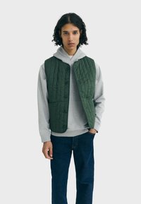 Grüner, gesteppter Weste mit Rundhalsausschnitt, Fronttaschen und Knopfverschluss, die über einem grauen Hoodie getragen wird, kombiniert mit blauen Jeans.