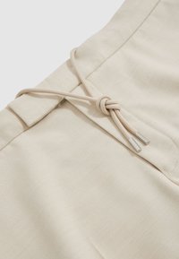 Ceinture en tissu beige avec un cordon noué doté d’embouts métalliques à chaque extrémité.